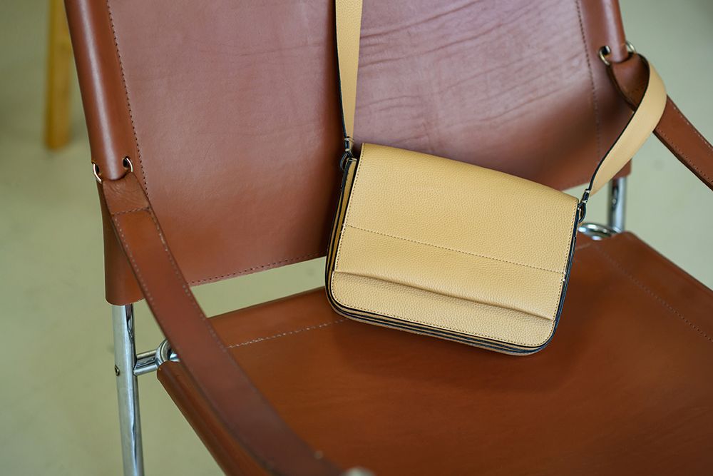 A beige leather crossbody bag sits on a brown vinyl chair against a pale green wall.