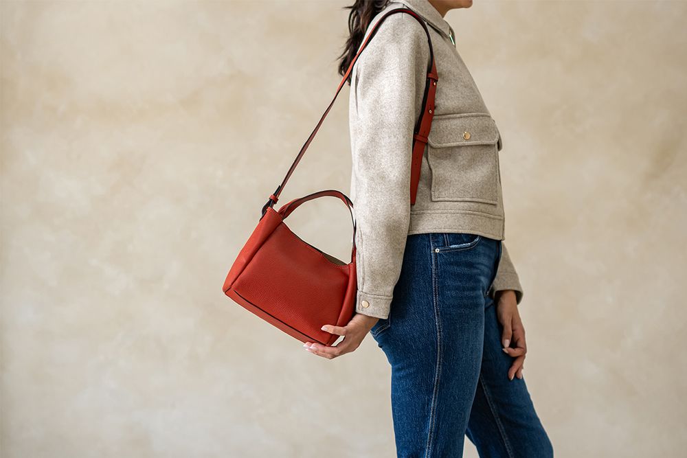 Woman in beige sweater and blue jeans carrying a red leather handbag in a studio setting.