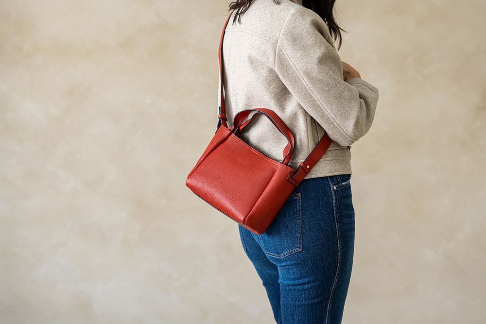 Woman wearing cream sweater and jeans with a stylish red leather handbag against neutral background.