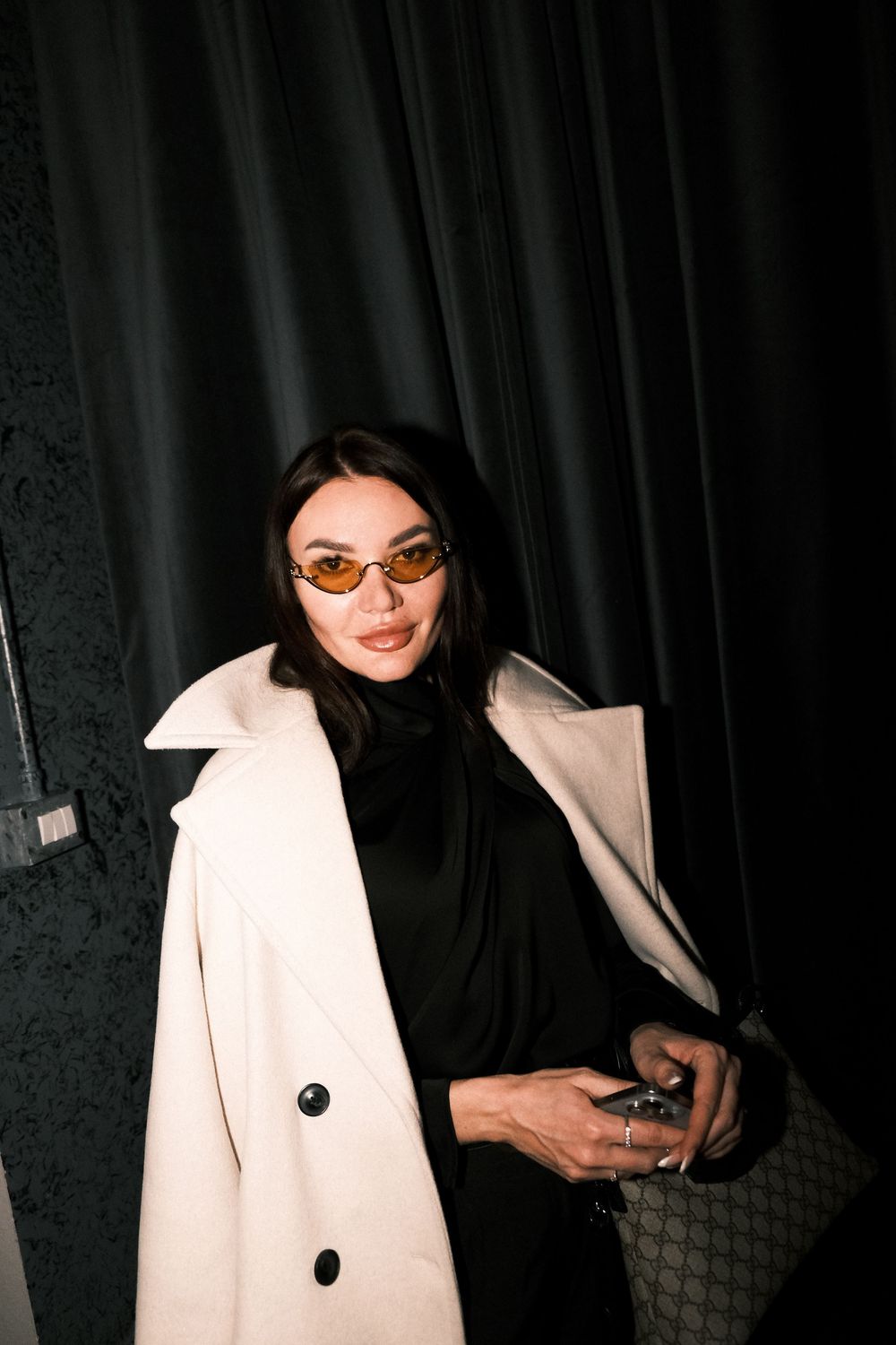 Stylish person wearing sunglasses, black turtleneck and cream coat poses against dark curtain in moody lighting.