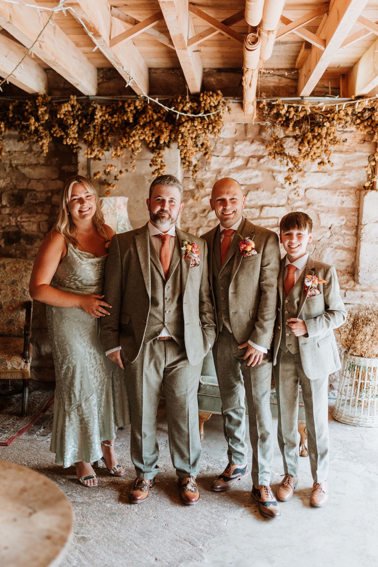 Wedding party in light gray suits poses together under rustic wooden beams decorated with dried flowers.