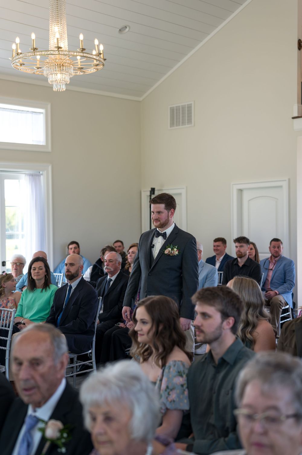 Wedding guests seated in an elegant white room with chandeliers during a ceremony.
