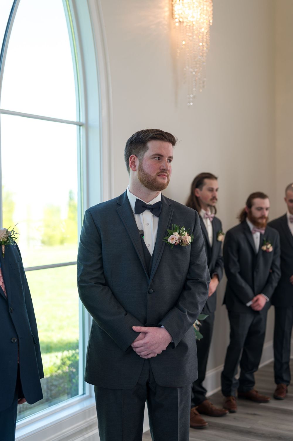 Groomsmen in black suits lined up against a bright window during a formal wedding ceremony.