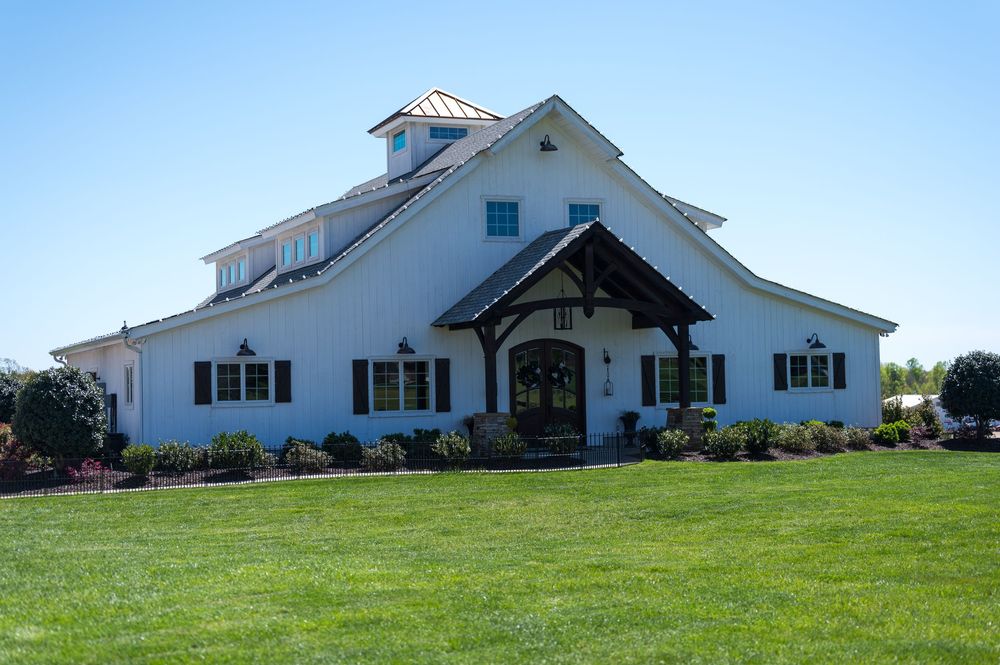 A white farmhouse with a gabled roof sits atop a green grassy hill against a bright blue sky.