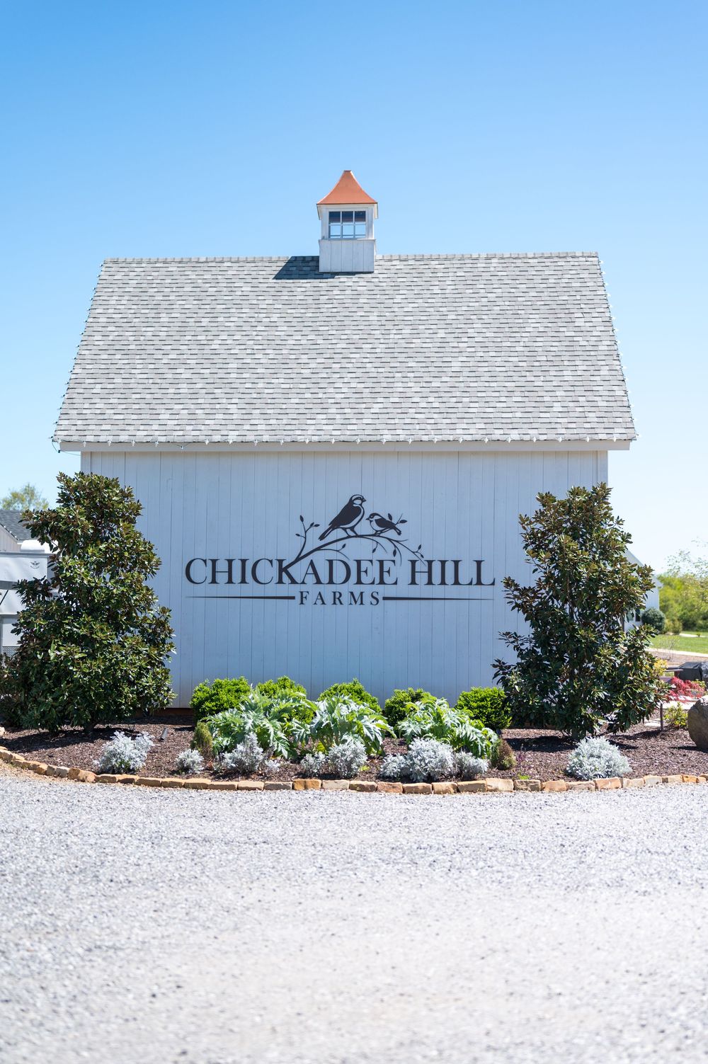 White barn with 'Chickadee Hill Farms' signage and landscaping against a bright blue sky.