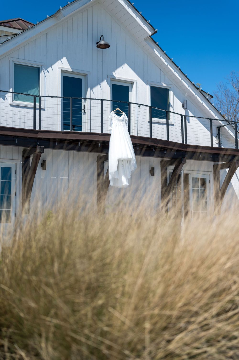 A white wedding dress hangs on display outside a white beach house with blue shutters and coastal grass in foreground.