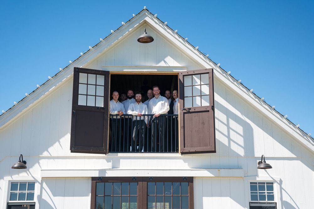 White farmhouse-style building with black windows and a decorative dormer balcony against bright blue sky.