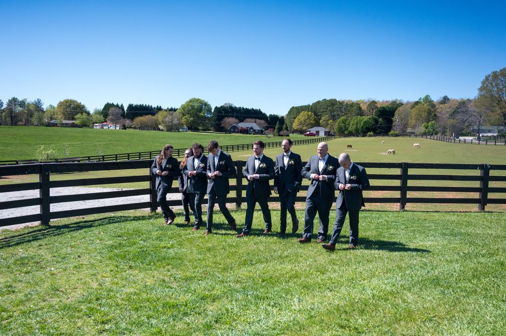 Wedding party members stand in line along a black and white fence on a sunny day at a countryside venue.