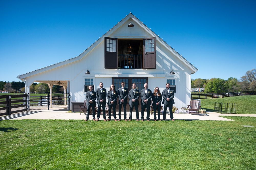 Wedding party standing in a row outside a white barn venue on a sunny day.