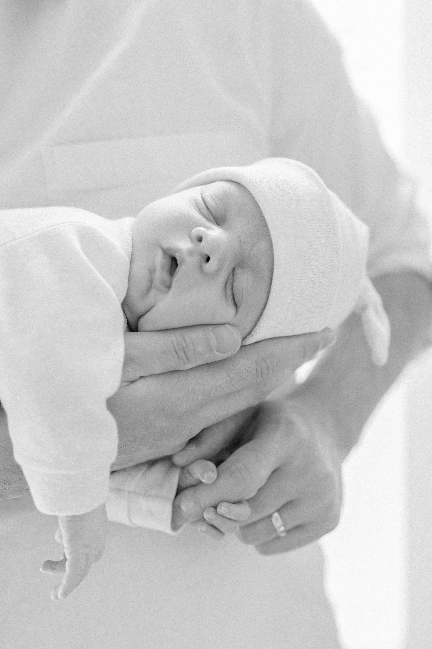 A newborn baby wrapped in white cloth is gently held in caring hands in a black and white photograph.