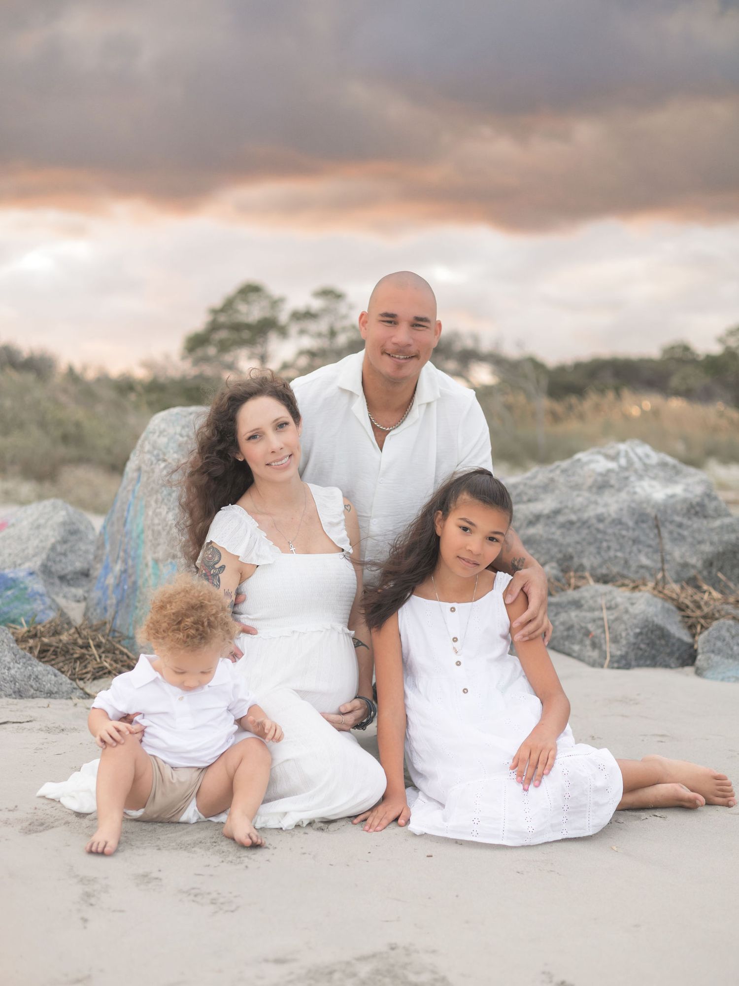 A family wearing white outfits poses together on a rocky beach at sunset creating intimate portraits.