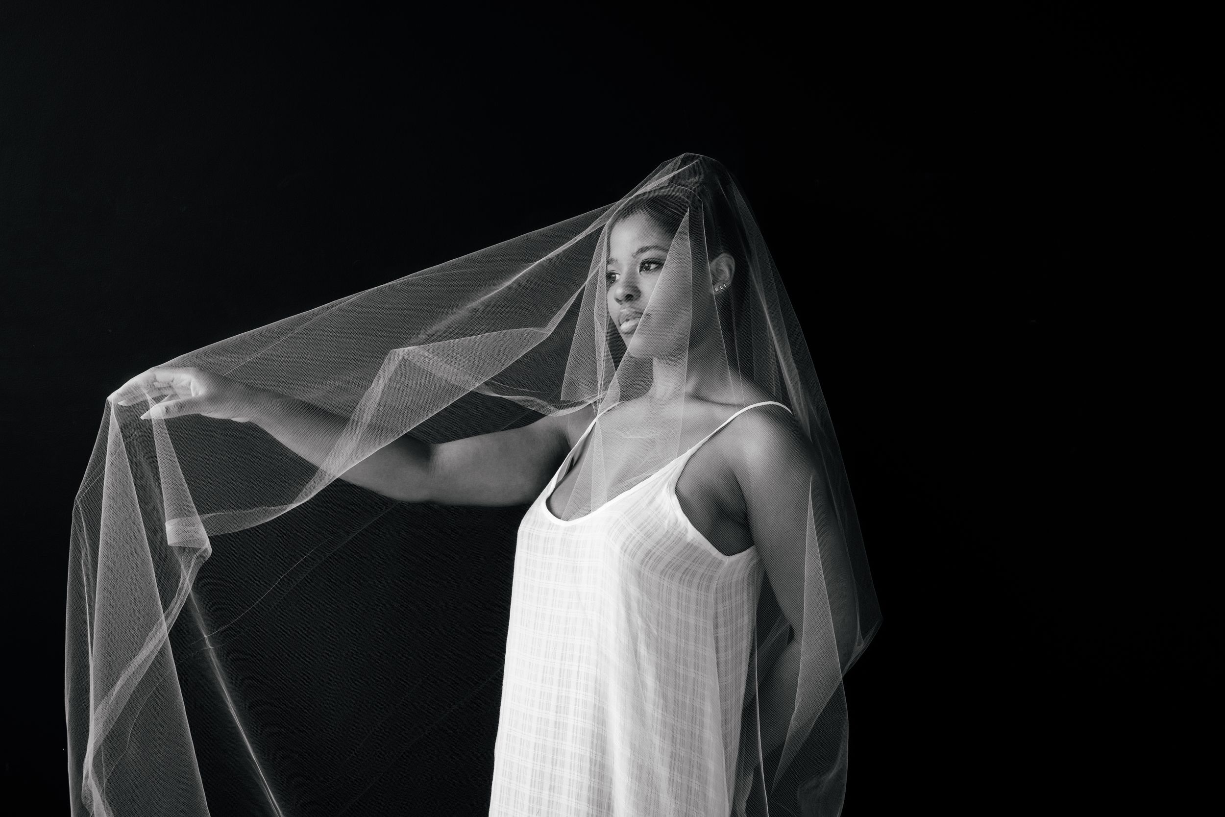 A dramatic black and white portrait of a figure in a white dress and flowing veil against a dark background.