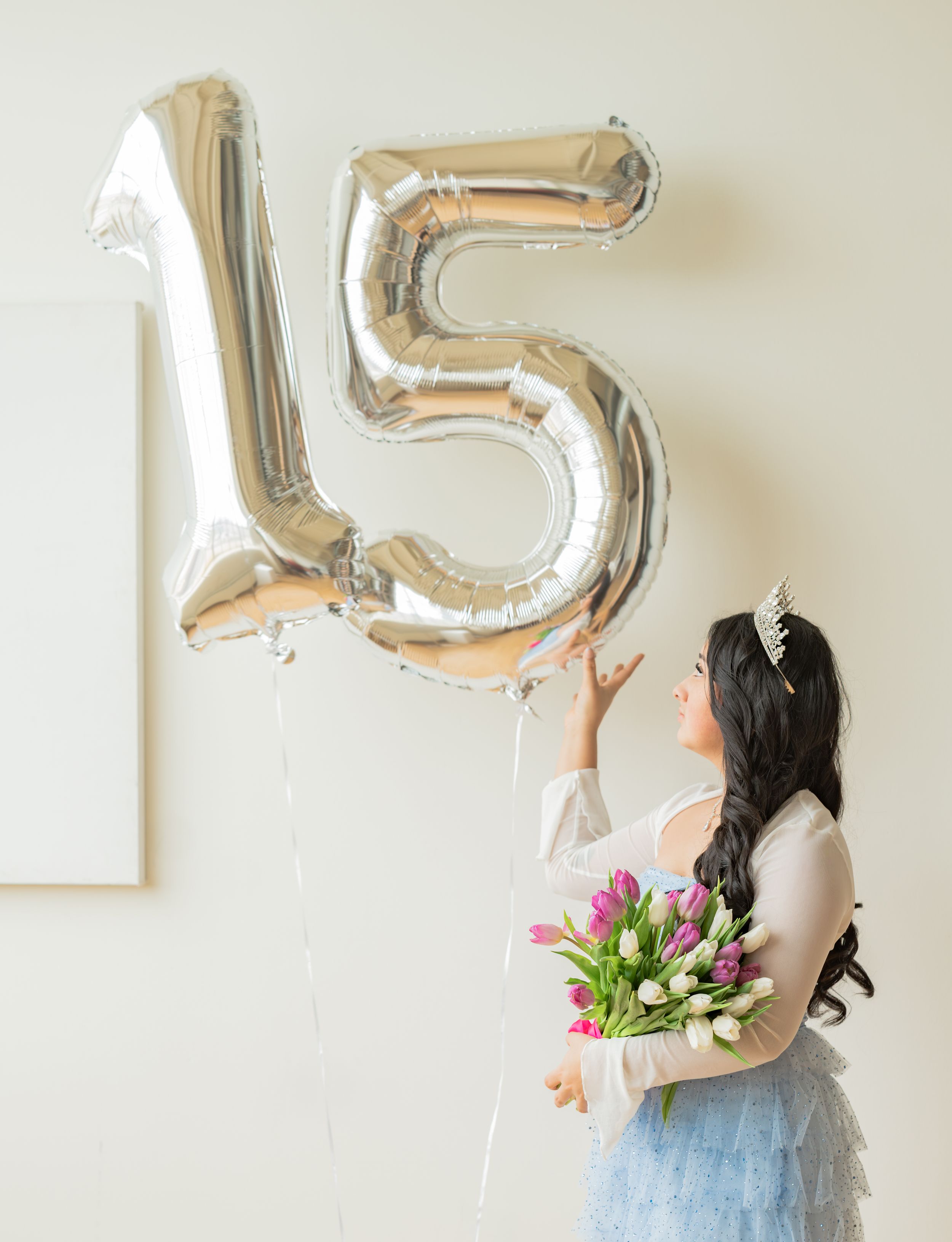 A young woman in a blue tulle skirt holds pink tulips beside silver number 15 balloons for a quinceañera celebration.