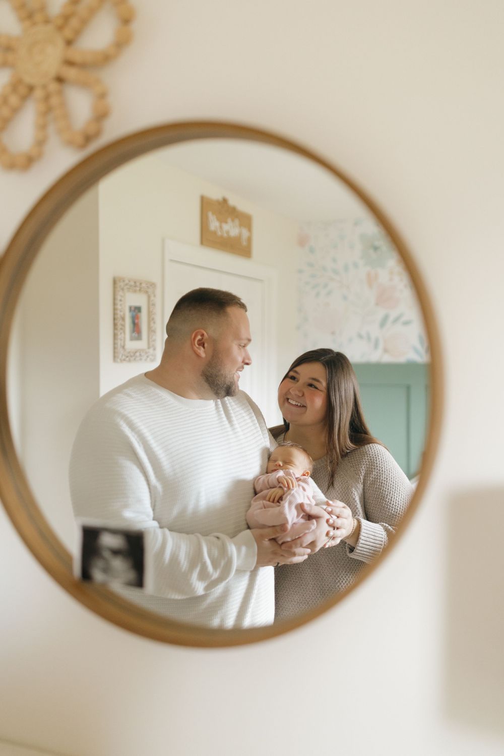 Family moment captured in oval mirror reflection showing parents holding their newborn.