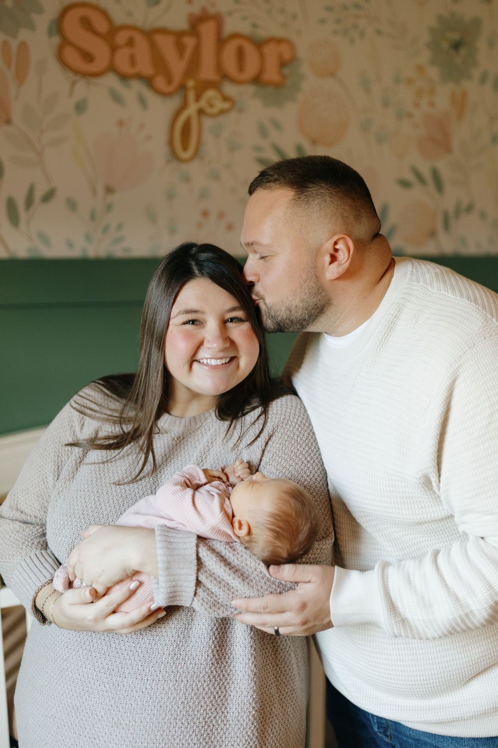A couple in matching cream sweaters holds their newborn baby in a nursery with floral wallpaper and name decor.