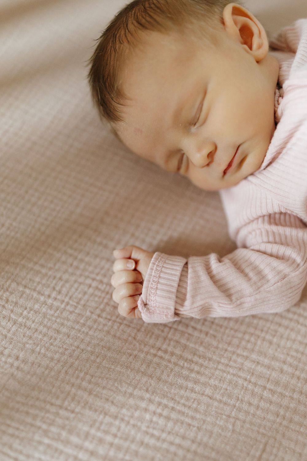 Peaceful sleeping baby wearing pink sweater rests on textured beige blanket.