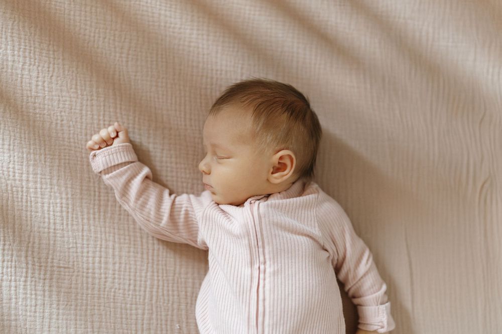 A sleeping baby wearing a white outfit lies on soft beige bedding in a peaceful pose.