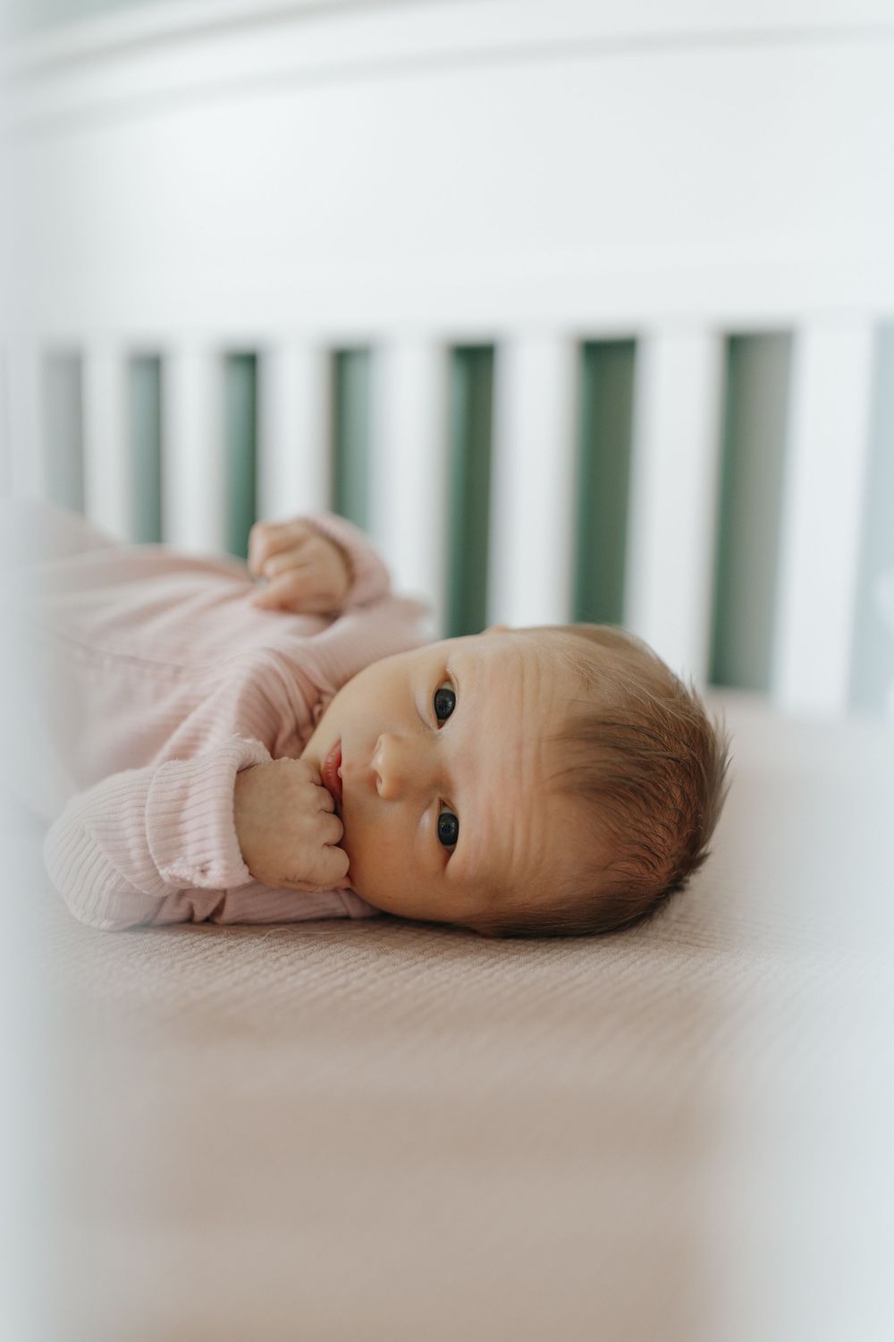 A newborn baby in pink sleepwear rests peacefully on white crib bedding.