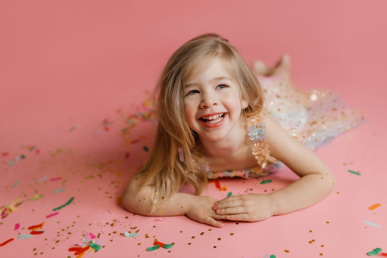 A person lies on pink background surrounded by colorful confetti and sprinkles creating a playful portrait series.