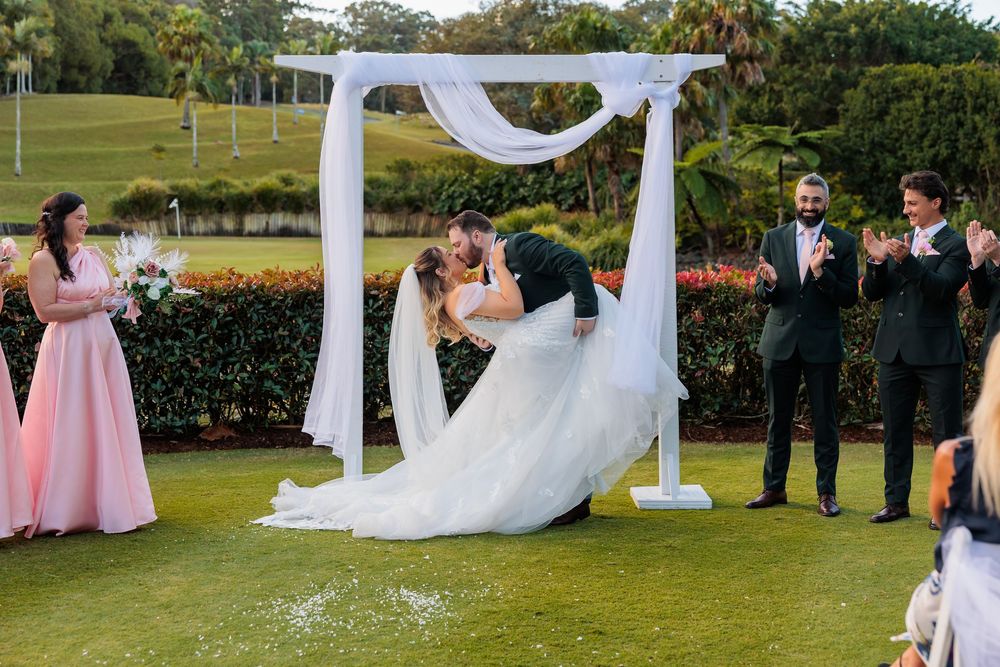 Bride and groom share first kiss on green golf course lawn with wedding guests applauding. Wedding Photography Coffs Harbour New South Wales