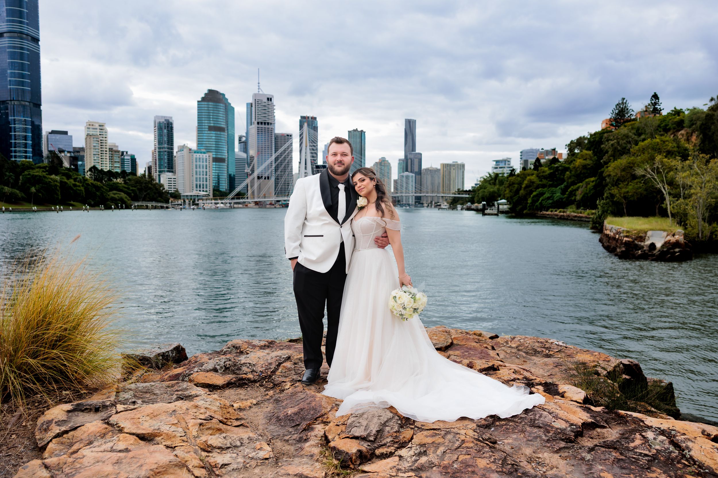 A couple poses on rocky outcrop with Brisbane cityscape and river in background.