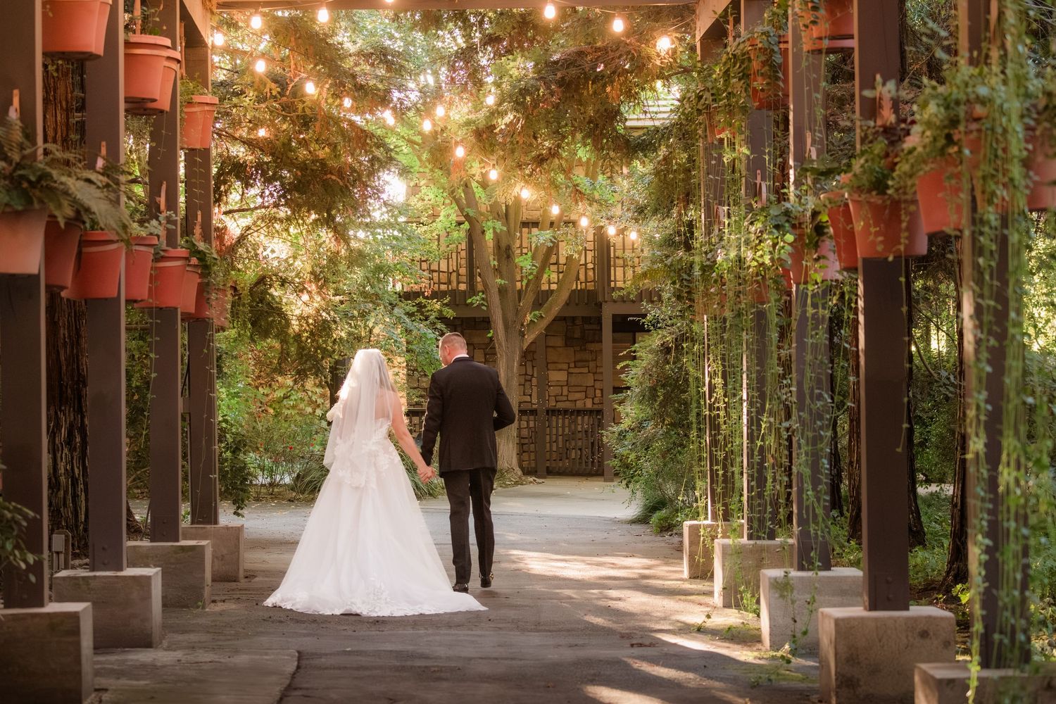 A wedding venue walkway with rustic wooden beams, string lights and hanging greenery creates a romantic atmosphere.