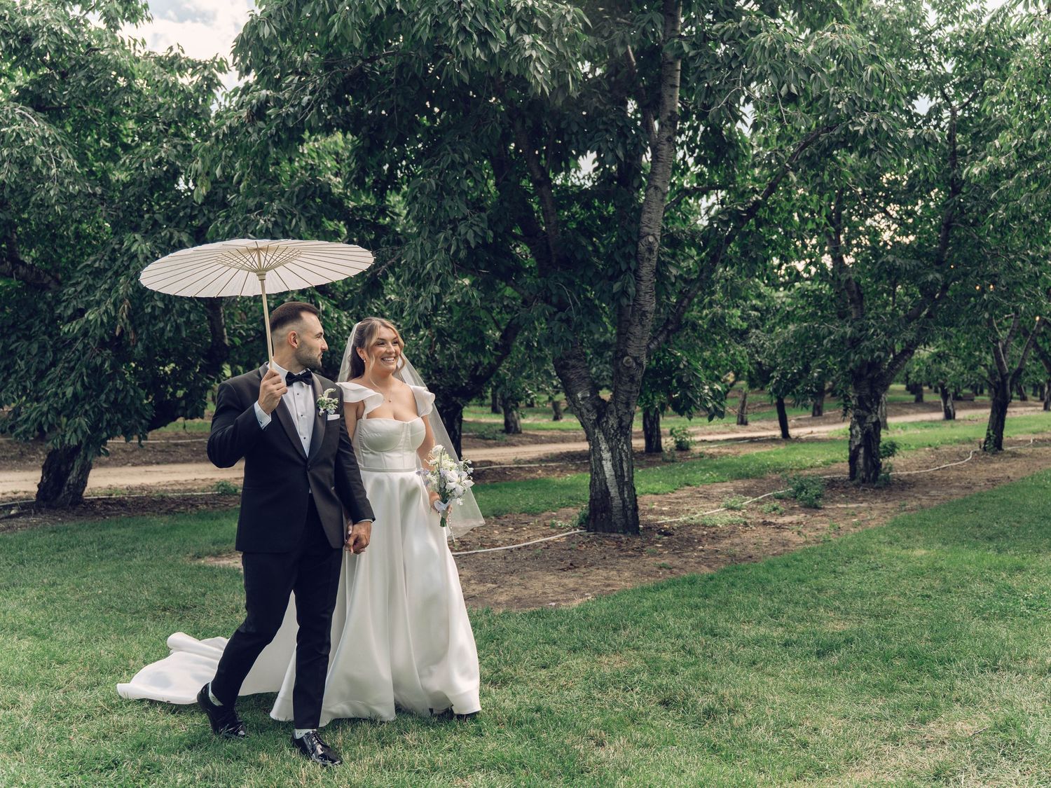 A couple in formal wedding attire walks together through a scenic garden setting with lush green trees and grass.