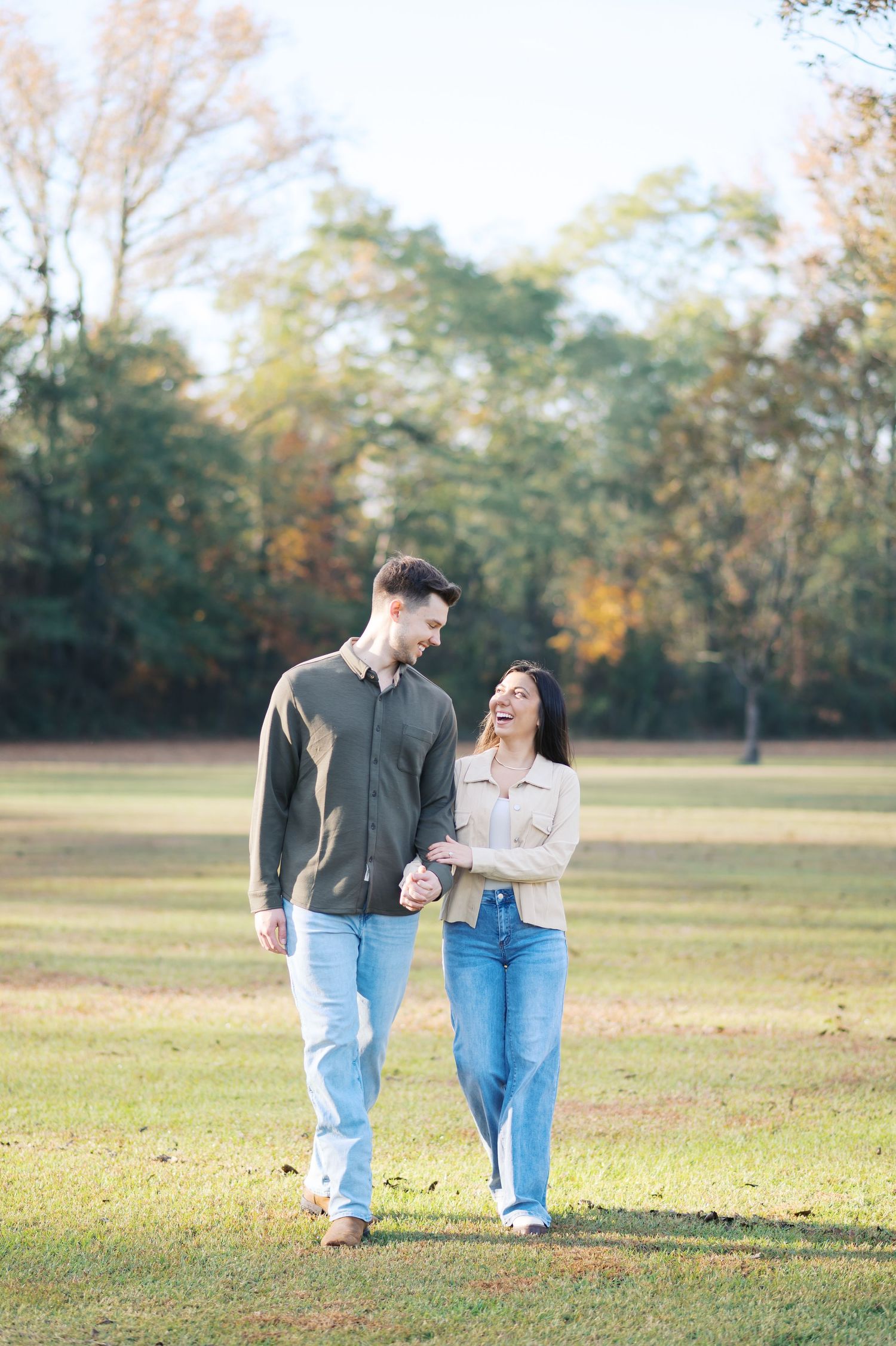 A couple walks hand in hand through a sunlit autumn field with golden trees in the background.