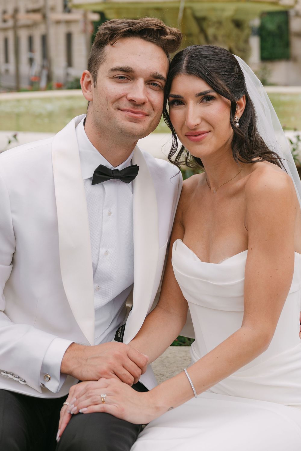 A bride in a white strapless gown and groom in white tuxedo jacket pose together outdoors on their wedding day.