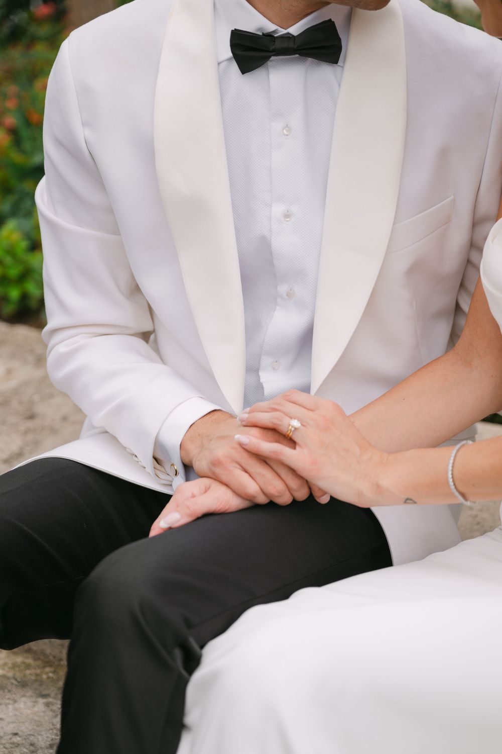 A couple in formal wedding attire sits together holding hands, with the groom in white tuxedo and black bow tie.