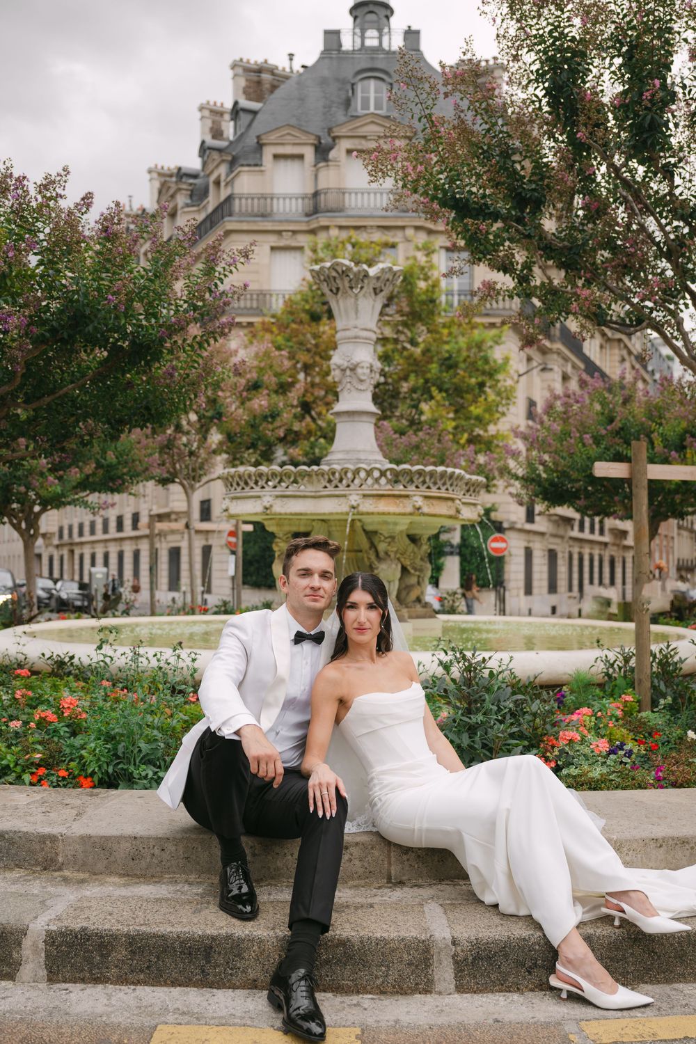 A couple in wedding attire sits together on stone steps in a romantic garden setting with blooming flowers.