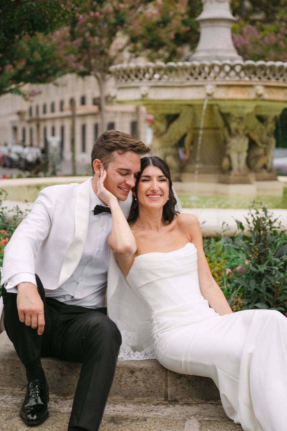A wedding couple shares a tender moment while sitting by an ornate fountain in an elegant garden setting.