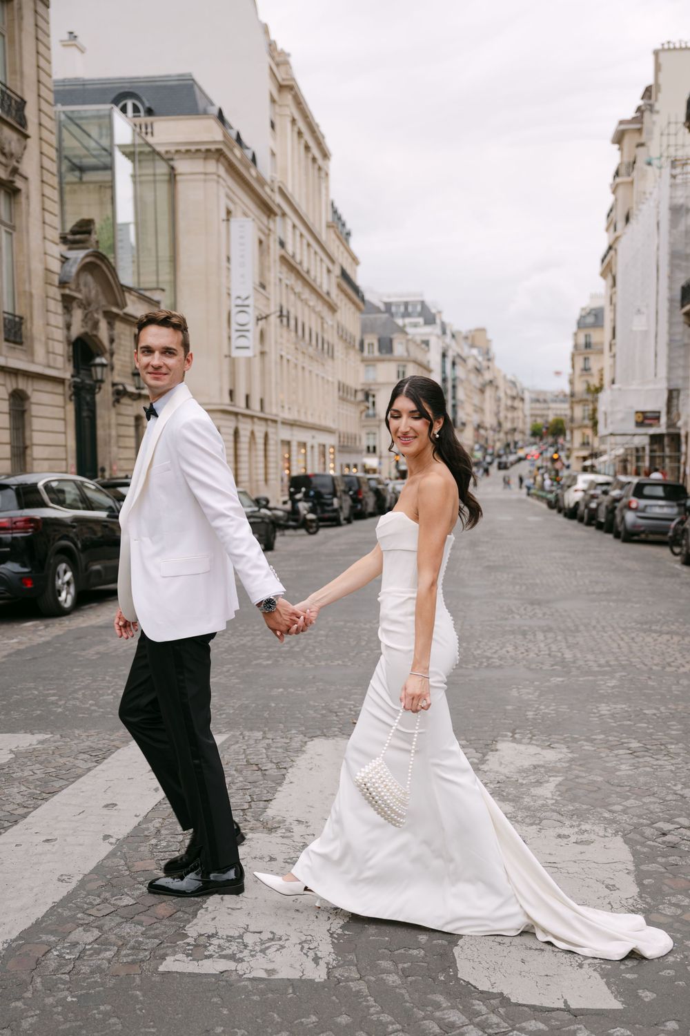 A couple in wedding attire dances playfully on a cobblestone street in Paris with elegant buildings in the background.
