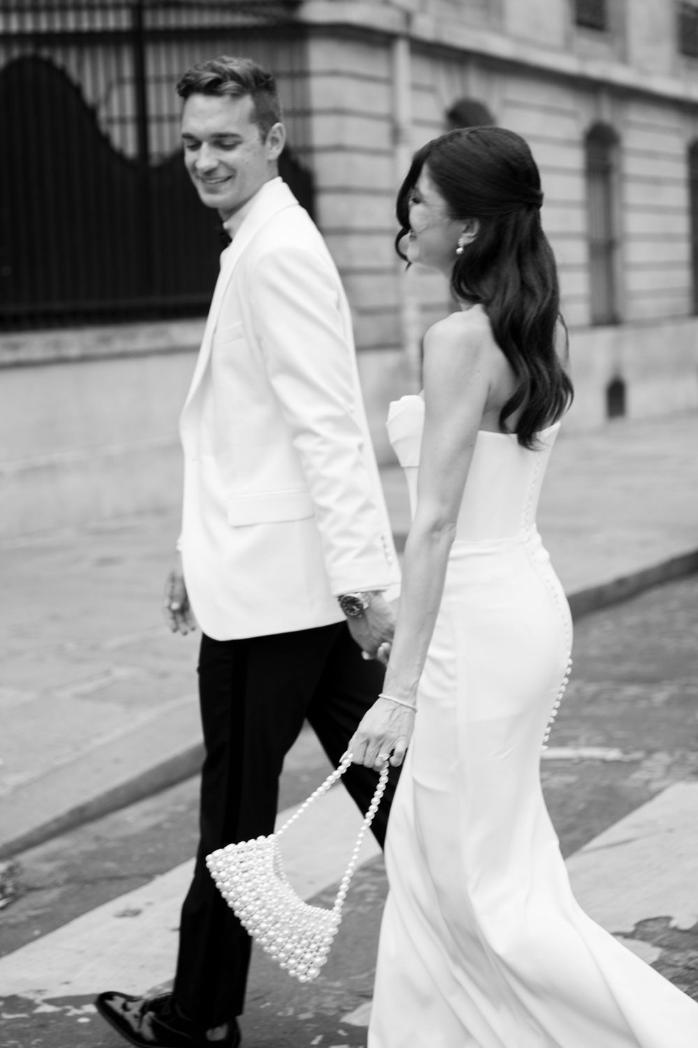 A couple in formal black and white attire walks together on a city street in a romantic black and white photograph.