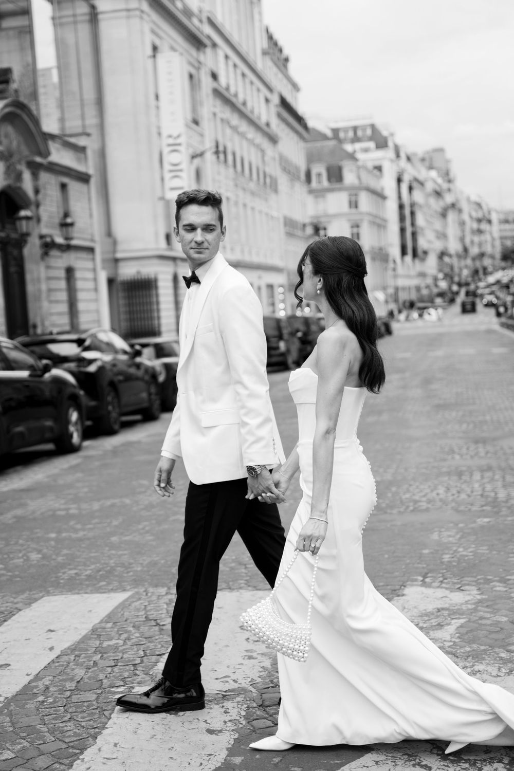 A bride and groom walk hand in hand on a Parisian street lined with classic buildings and parked cars.