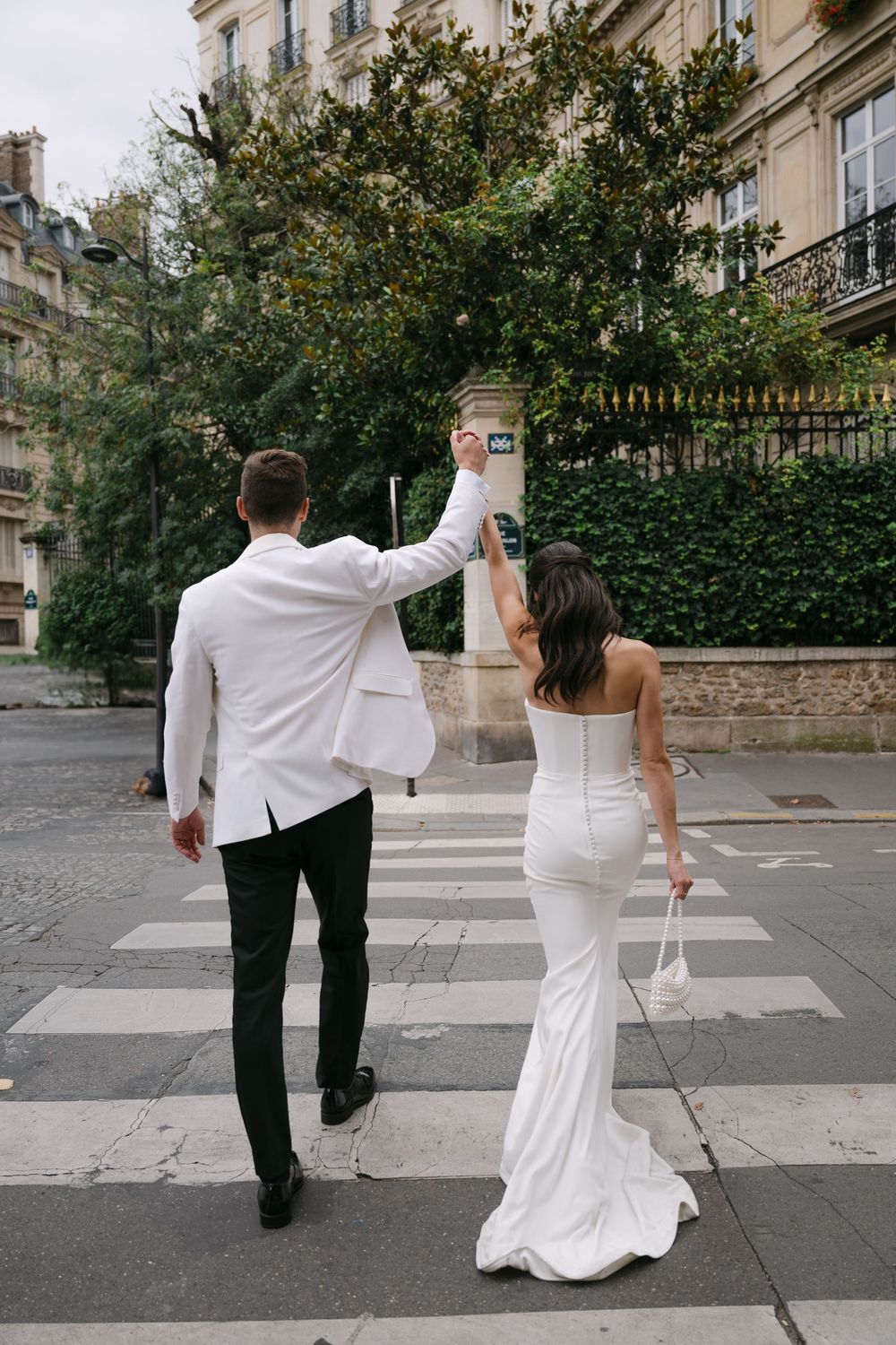 A couple in wedding attire walking across a crosswalk in a charming European neighborhood with stone buildings.