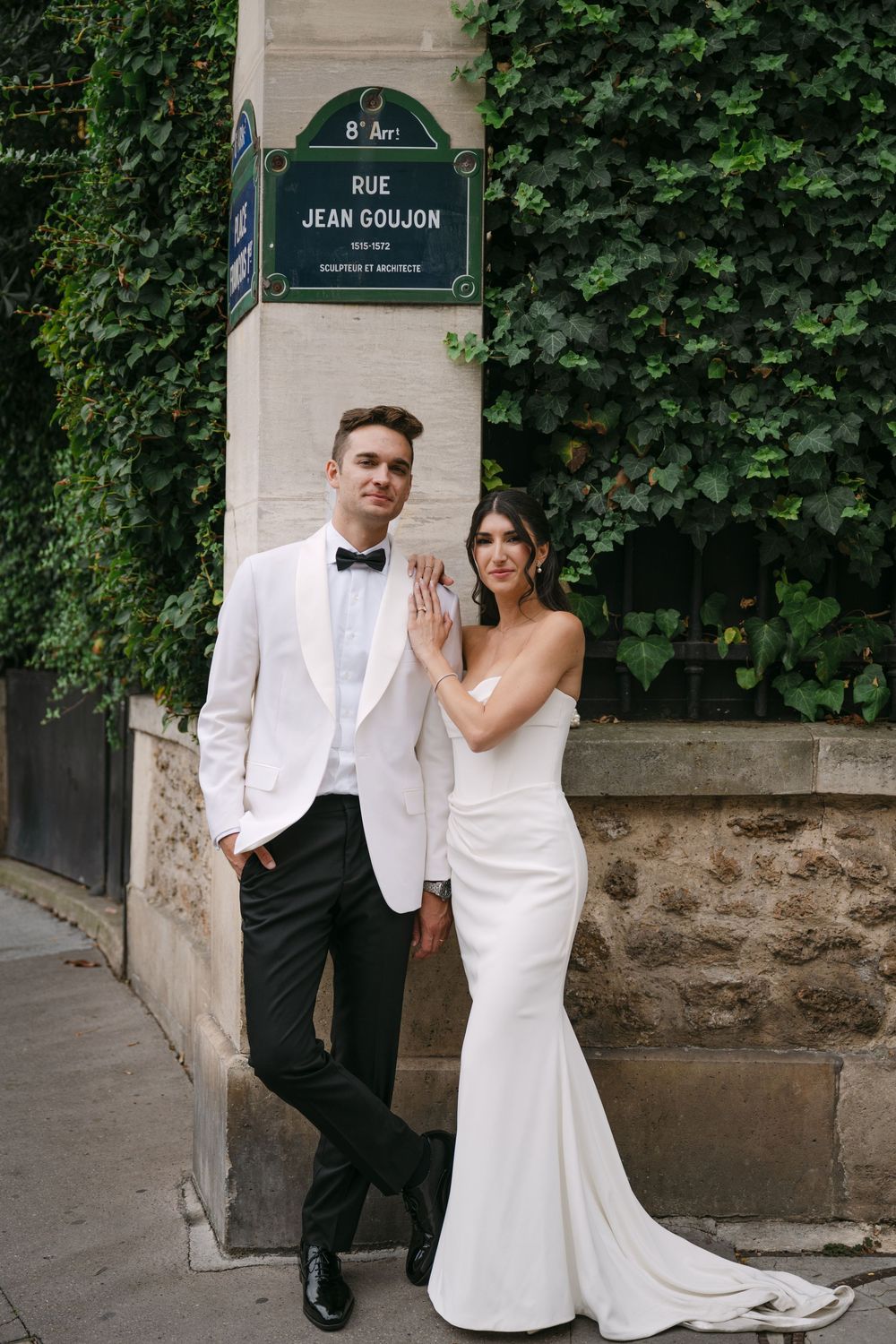 A bride and groom pose together on a Paris street corner with green ivy and street signs visible in the background.