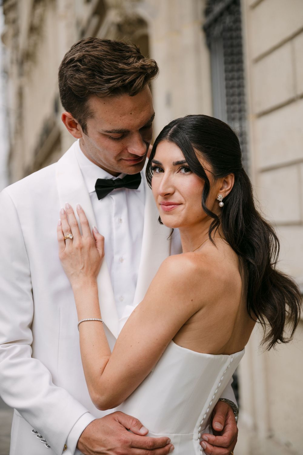 A romantic wedding portrait of a couple in white formal attire against a classic architectural backdrop.