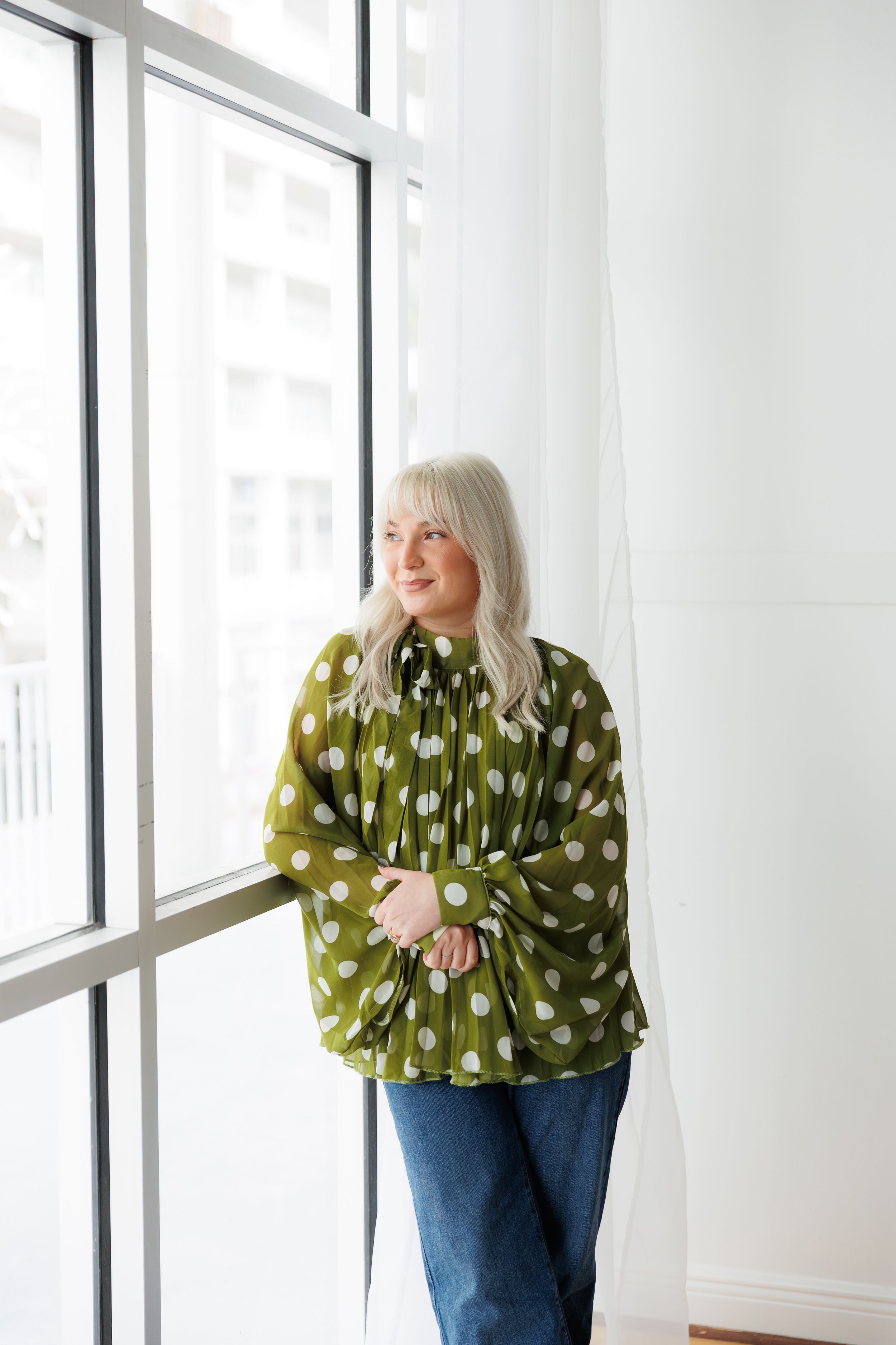Person in olive green polka dot sweater and jeans poses near bright windows in white minimalist space.