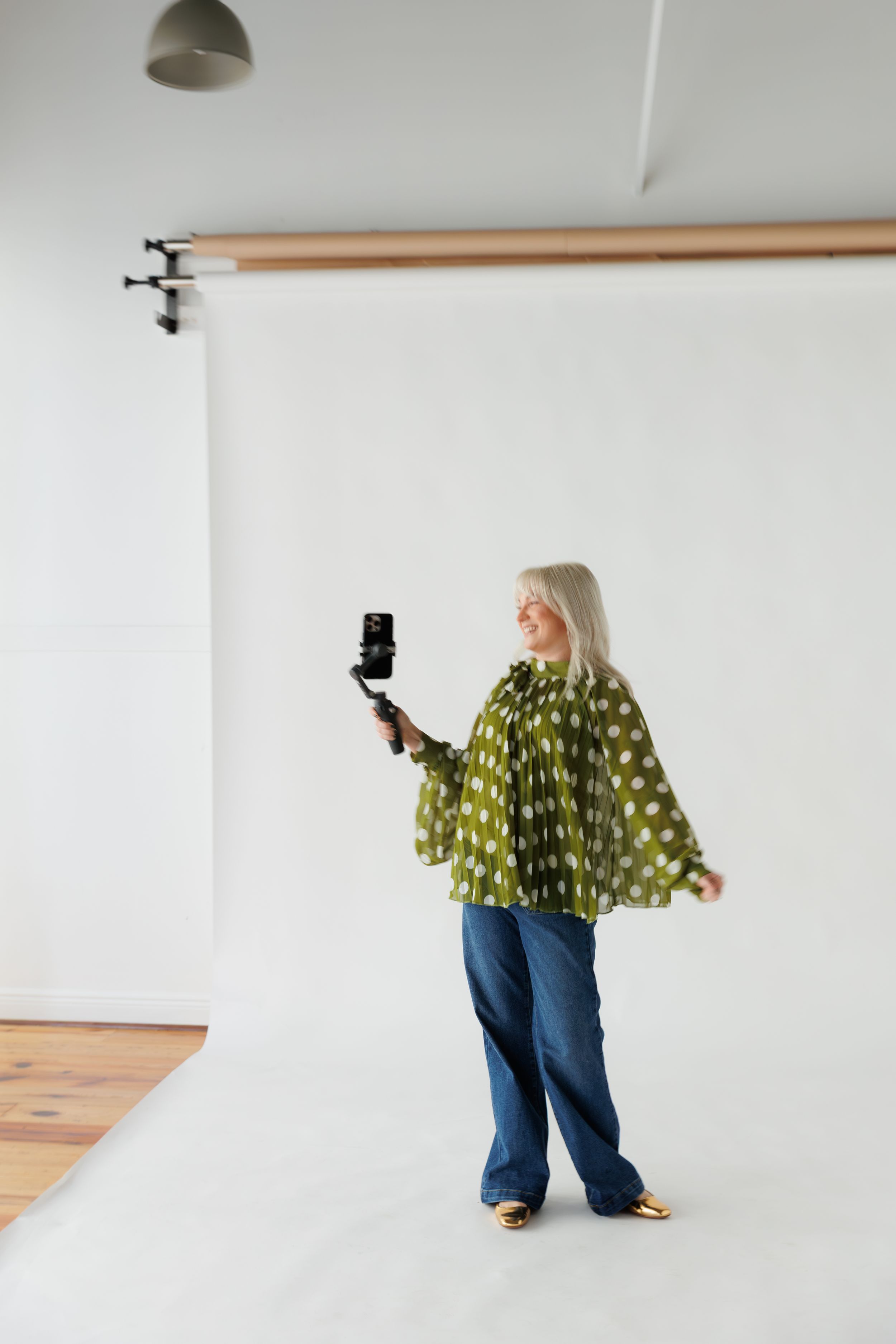 A person wearing a green polka dot top and blue jeans poses playfully in a white studio setting.