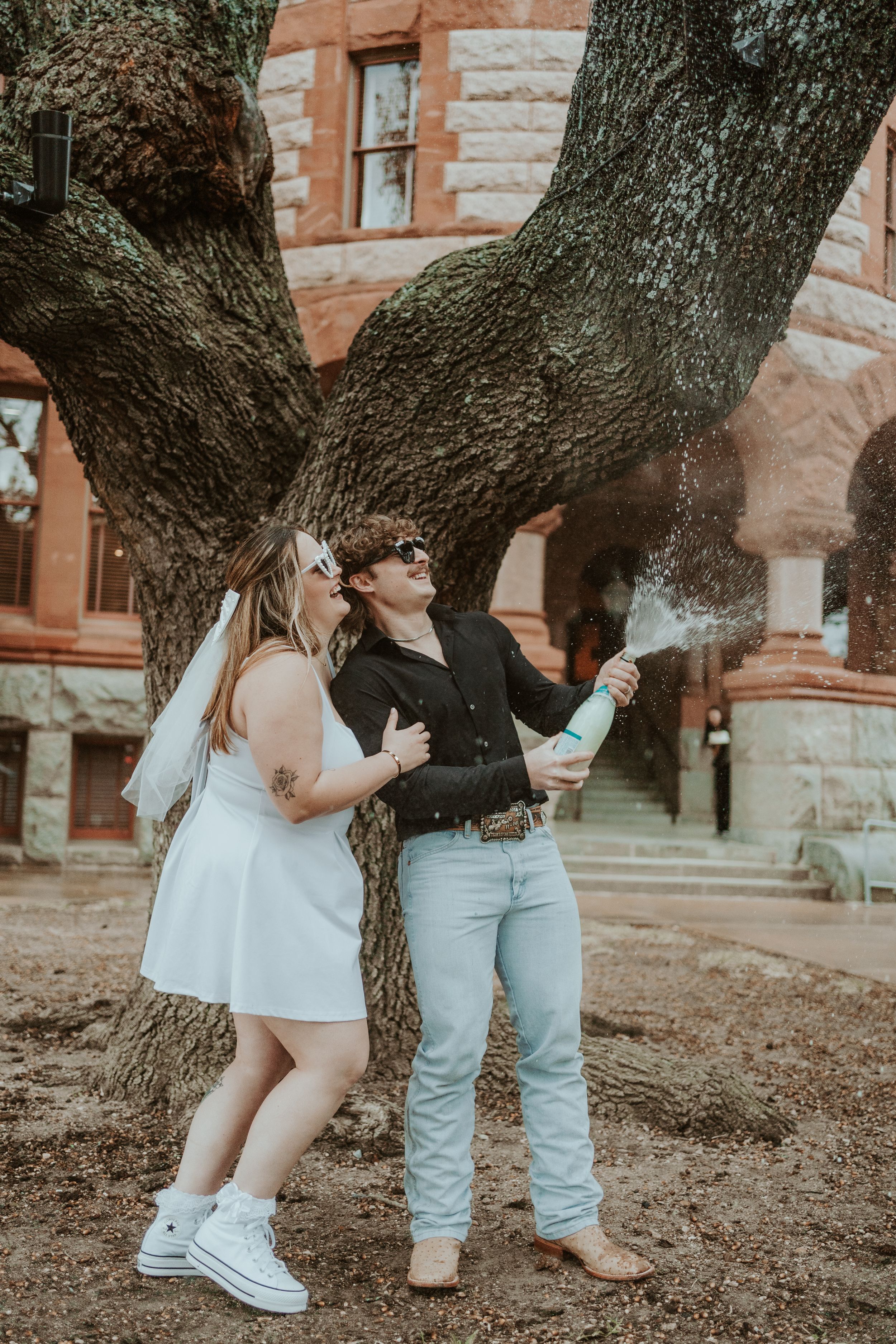 Couple's feet visible beneath a large tree trunk against a brick building backdrop.