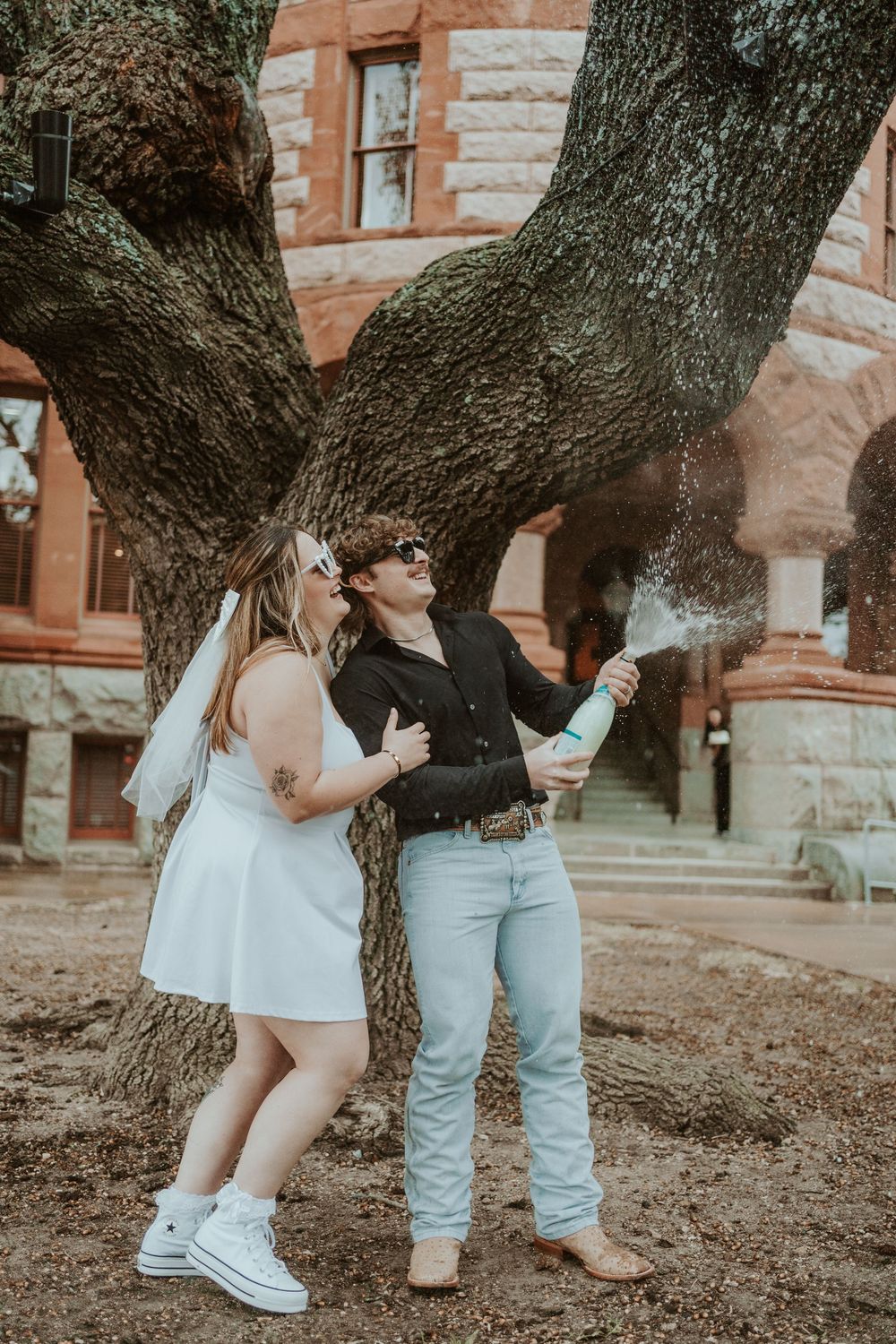 Couple's feet visible beneath a large tree trunk against a brick building backdrop.
