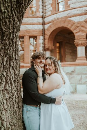 Romantic couple embrace under a tree in front of historic stone building.