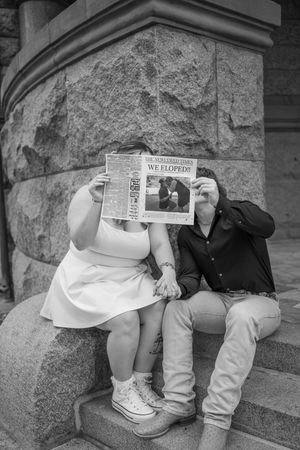Couple playfully hiding behind newspapers while sitting on stone steps.