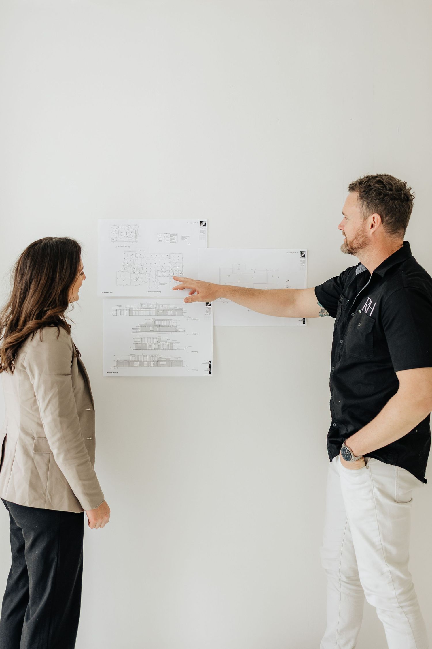 Business colleagues point and discuss plans posted on white wall during collaborative meeting.