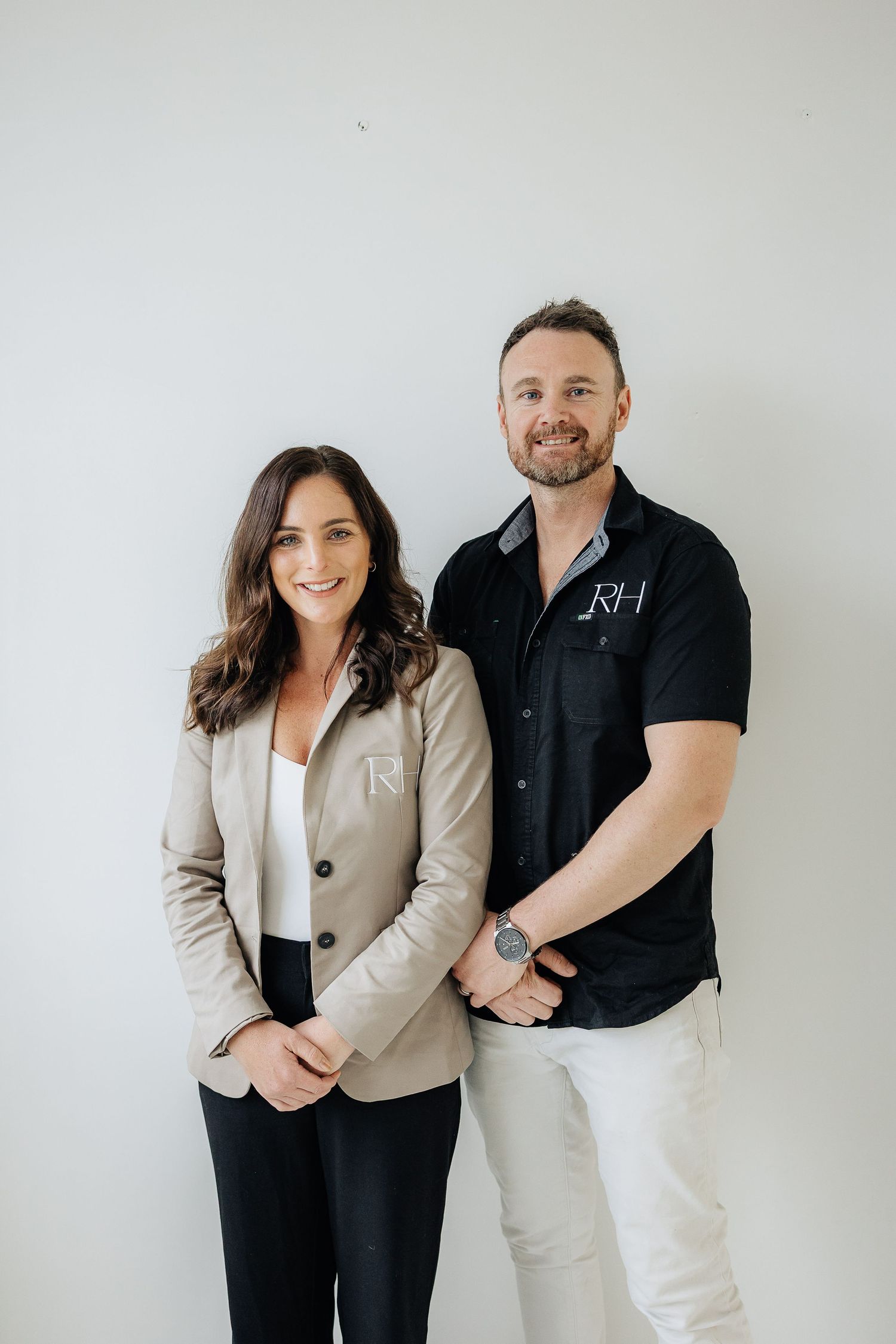 Professional business partners in contrasting black and beige outfits pose against white wall in studio setting.