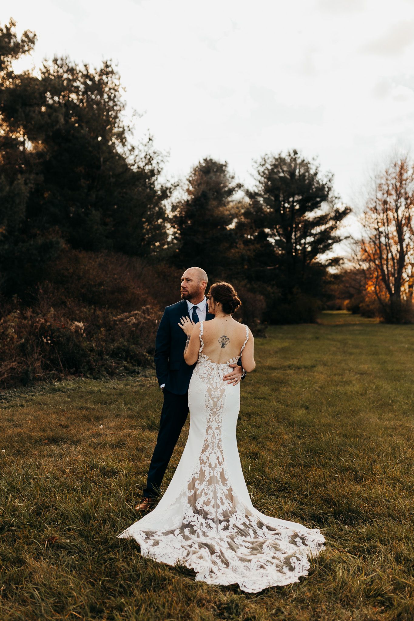 A couple in wedding attire embraces during sunset in a grassy field with trees in the background.