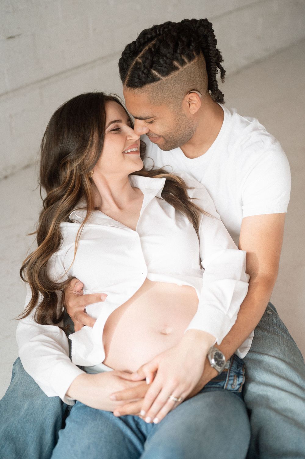 Intimate maternity photo of couple in white shirts embracing tenderly against neutral wall background.