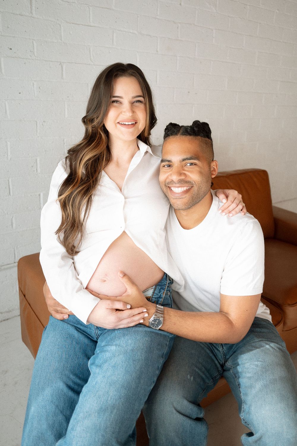 A couple in casual white tops and jeans sit together on a brown leather couch against a white brick wall.
