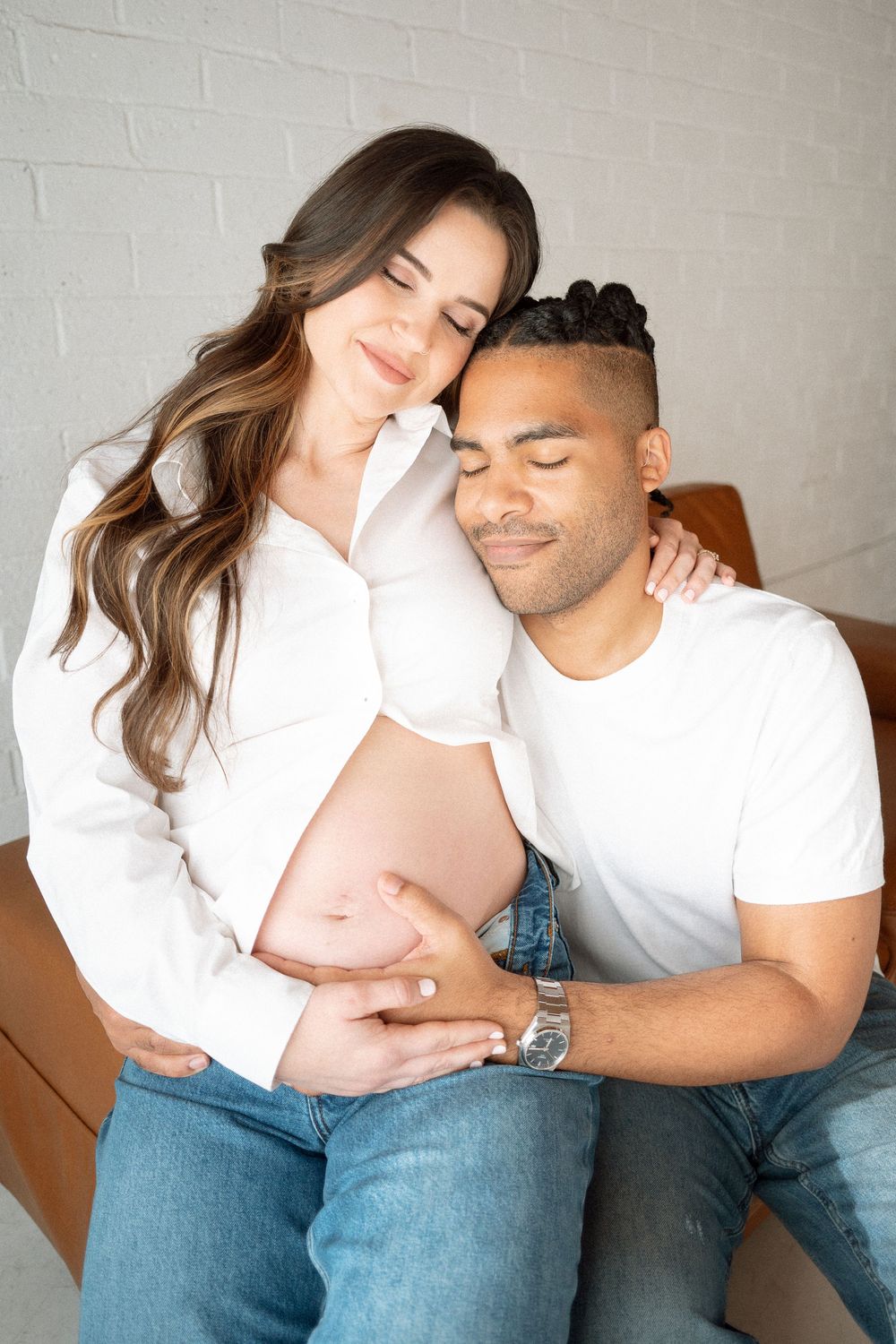 A couple in matching white shirts and blue jeans share intimate moments while sitting together on a couch.